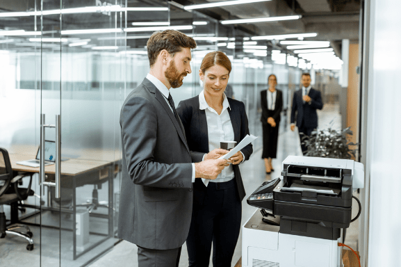 Two professionals in an office setting observe printed pages at a copier, as it is meant to represent the importance of Managed Print Services.