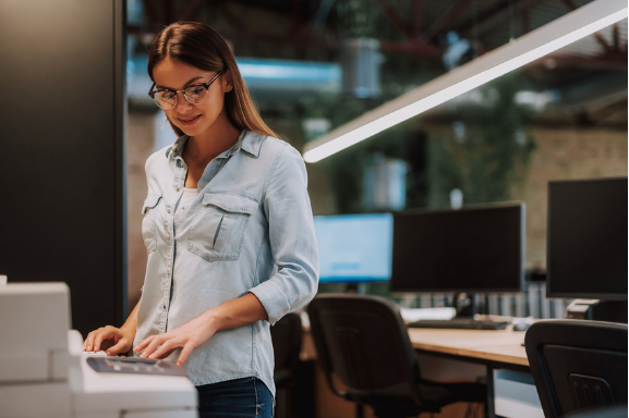 A young professional woman stands next to an office copier representing output management.