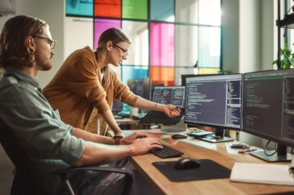 Two young professionals review code on one of three computer screens signifying IT Services