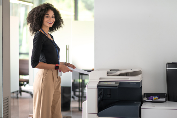 A professional woman with curly hair smiles as she collects documents from a multifunction printer in a bright, modern office.