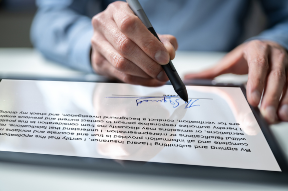 Close-up view of a person's hands using a stylus to provide an electronic signature on a digital tablet. The screen displays the signature in blue ink, along with lines of blurred text in the background, emphasizing the process of digitally signing documents.