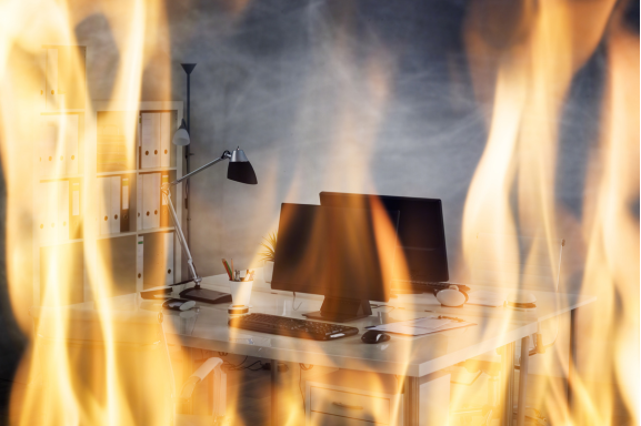 A computer workstation engulfed in flames, with smoke rising around the room, illustrating the urgent need for disaster recovery in an office setting.