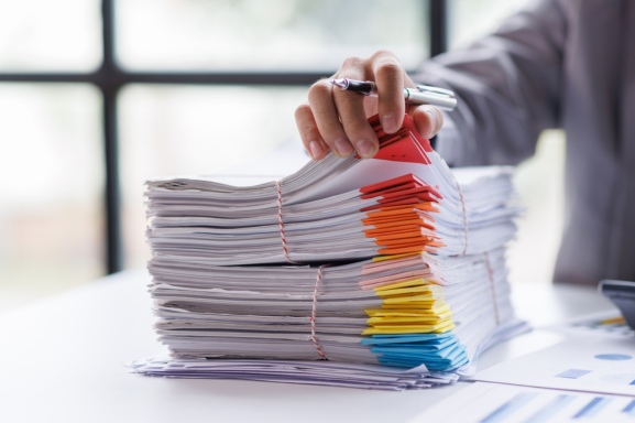 A person's hand rests on top of a large stack of assorted documents with colorful tabs, illustrating the high output typical of production print services in an office environment with natural light.