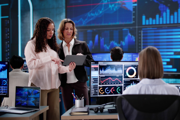 Two women stand in a high-tech office reviewing a tablet while others look at data on their screens signifying the importance of quality IT support for modern businesses.