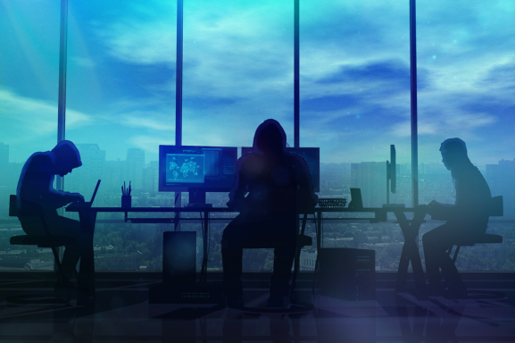 Three nefarious, hooded figures sit at a desk in a high-rise city building. symbolizing the need for cybersecurity.