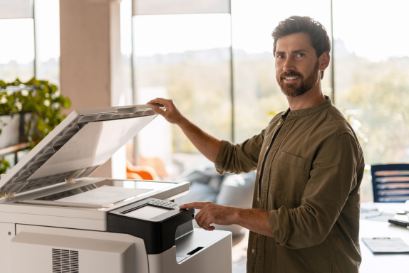 A young professional stands at a multifunction office copier in a bright, modern office.