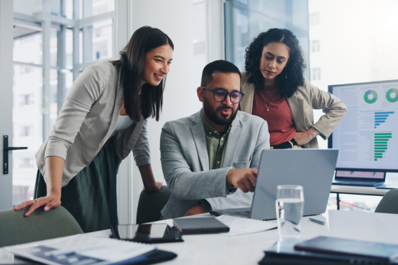 A group of young professionals stand around a laptop with a screen of charts and graphs in the background signifying Managed IT Services.