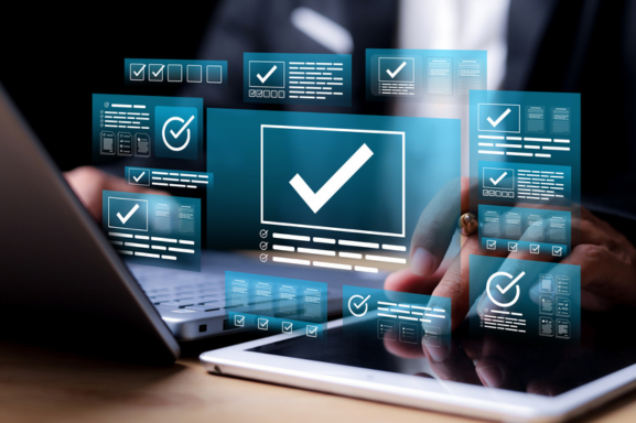 Close up of a professional working on a laptop with a tablet with icons of windows and checks above it as a conceptual image of IT compliance.