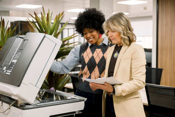 Two professional women stand in front of a multifunction office printer discussing outputs smiling.